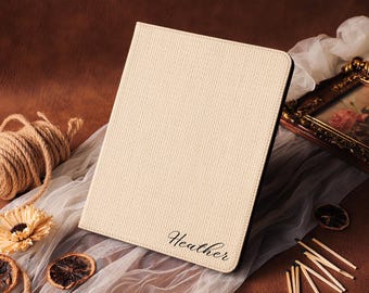 May include: Cream-colored tablet case with the name "Heather" in cursive script. The case has a textured surface and a black border. The background includes a roll of twine, dried flowers, and a framed picture.