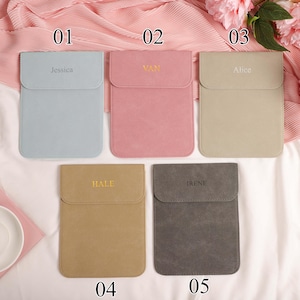 May include: Five personalised laptop sleeves in pastel shades: light blue, pink, beige, tan, and grey. Each sleeve features a flap closure and an embroidered name: Jessica, Van, Alice, Hale, and Irene. The sleeves are arranged on a pink fabric.