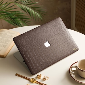 Peut inclure: Une housse d'ordinateur portable marron foncé avec un motif texturé en peau de crocodile et une découpe pour le logo Apple. La housse présente les initiales MA dorées en relief dans le coin inférieur droit. L'ordinateur est fermé.
