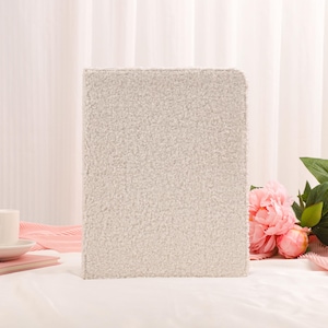 May include: A rectangular, light gray, textured book cover stands upright. A white teacup and saucer sit on a pink book, with pink peonies and fabric in the background. The cover has a soft, plush appearance.