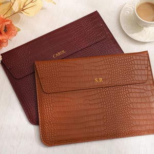 Könnte beinhalten: Zwei Laptop-Hüllen mit Krokodil-Textur. Eine ist weinrot, die andere warmbraun. Die weinrote Hülle hat den Namen "CAROL" in Gold geprägt, die braune Hülle die Initialen "S.R.".