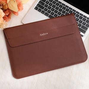 Könnte beinhalten: Eine braune Laptop-Hülle aus Leder mit dem eingeprägten Namen "Andrew" auf der Vorderseite. Die Hülle ist rechteckig mit einem Klappenverschluss. Ein Laptop, eine Sonnenbrille und Blumen sind im Hintergrund.