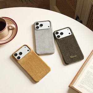 May include: Three rectangular smartphone cases with a soft, textured finish in mustard yellow, light gray, and olive green. Each case features a camera cutout and a small, embroidered name in cursive script: Howard, Harrison, and Charles.