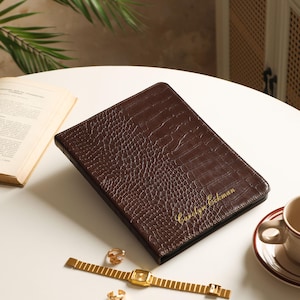 Puede incluir: Una carpeta rectangular de color marrón oscuro con textura de piel de cocodrilo y el nombre Carolyn Eckman impreso en color dorado en la portada. La carpeta se acompaña de un reloj de pulsera de metal dorado y dos anillos dorados.