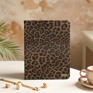May include: A rectangular notebook cover featuring a brown and black leopard print pattern. The bottom right corner displays the gold-colored initials HD. The cover has a smooth texture and rounded top corners, standing upright on a white surface.