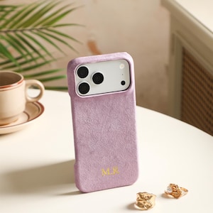 May include: A lavender-colored phone case with a soft, textured finish and a gold-colored monogram reading M.K. at the bottom. The case features a rectangular cutout for the camera lenses and is positioned upright on a smooth surface.
