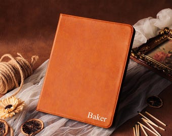 May include: A leather-bound, orange-colored portfolio with the name "Baker" embossed in white. The portfolio is rectangular and appears to be closed. The background includes decorative elements like a framed picture and dried citrus slices.