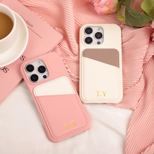 May include: Two leather phone cases, one pink and one cream, with a pocket design. The pink case has the initials "L.S." and the cream case has "T.Y." in gold lettering. A cup of coffee and a pink book are also in the image.