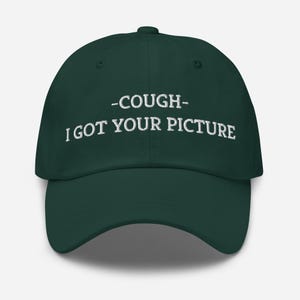 May include: A dark green baseball cap with white embroidered text that reads " -COUGH- I GOT YOUR PICTURE".