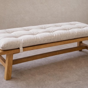 May include: A wooden bench with a light-colored, tufted cushion. The bench has a rectangular shape with a natural wood finish. The cushion is secured with white straps. The design is simple and modern, suitable for various interior styles.
