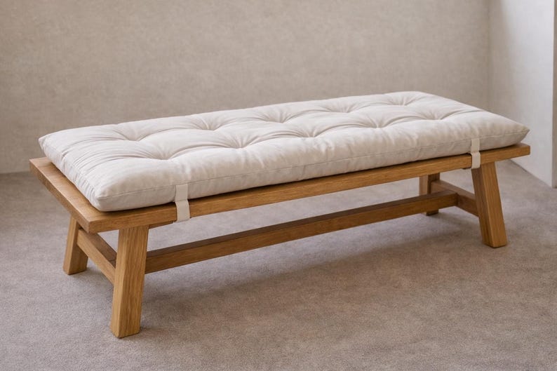 May include: Wooden bench with a cream-coloured, buttoned cushion. The bench has a rectangular shape and a natural wood finish. The cushion is secured with white straps. The design is simple and modern, suitable for various interior styles.