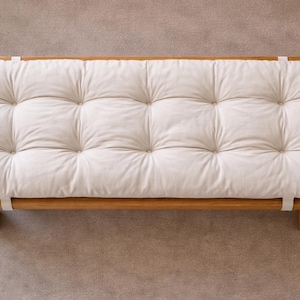 May include: A wooden platform bed frame with a tufted, off-white cushion. The cushion is rectangular and has a grid pattern of stitched squares. White straps secure the cushion to the frame. The bed frame is made of light-coloured wood.