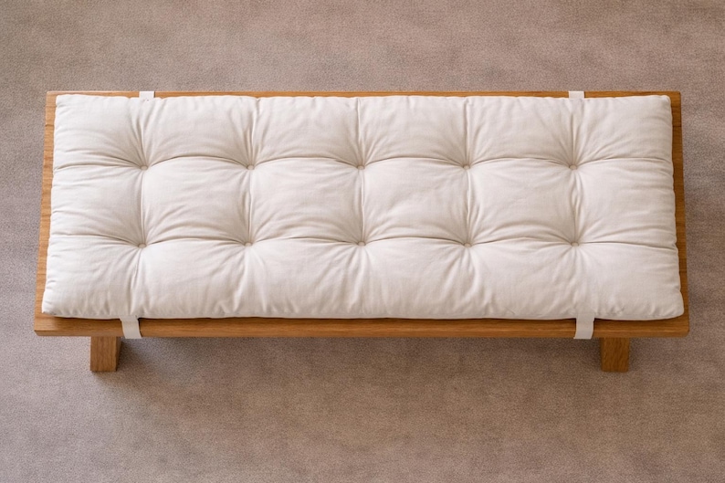 May include: A wooden platform bed frame with a tufted, off-white cushion. The cushion is rectangular and has a grid pattern of stitched squares. White straps secure the cushion to the frame. The bed frame is made of light-coloured wood.