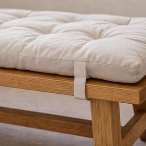 May include: A cream-coloured, quilted cushion rests on a wooden bench. The cushion is rectangular and made of a soft fabric. The bench is constructed from light-coloured wood, showing the wood grain.