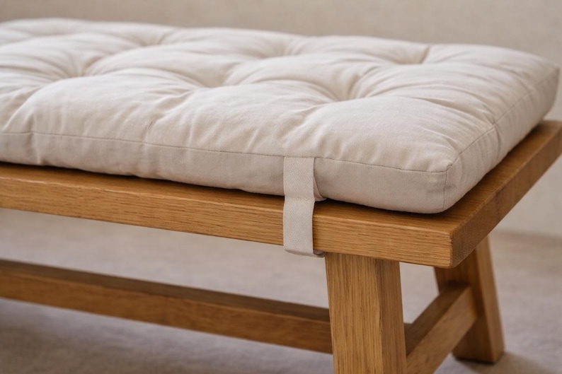 May include: A cream-coloured, quilted cushion rests on a wooden bench. The cushion is rectangular and made of a soft fabric. The bench is constructed from light-coloured wood, showing the wood grain.