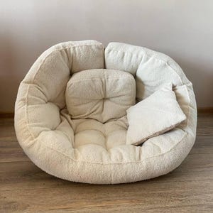May include: A cream-coloured, round, plush chair with a matching square cushion and a smaller, rectangular cushion. The chair has a tufted design and is made of a soft, textured fabric. The cushions are also made of the same material.
