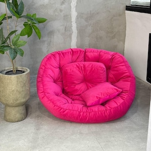 Op de afbeelding: Een felroze vloerkussen, rond van vorm, met een vierkant en een rechthoekig kussen, allemaal in dezelfde kleur. Het kussen staat op een grijze vloer, met een plant in een pot en een grijze muur op de achtergrond.