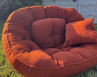 May include: A round, oversized floor chair upholstered in a textured, rust-orange sherpa fabric. The chair features a deep, tufted seat and backrest with a matching square throw pillow resting on the right side, creating a soft, plush appearance.