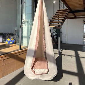 以下が含まれることがあります： オフホワイトの布地で作られたハンギングポッドチェア。前面の開口部にはダスティローズピンクの縁取りが施され、底面には同色の円形クッションが配置されています。構造は円錐形で上部に向かって細くなっており、広い入り口があります。
