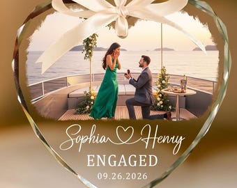 May include: A heart-shaped glass ornament featuring a printed photograph of a marriage proposal on a boat deck. The text reads Sophia and Henry, ENGAGED, and 09.26.2026. A white satin ribbon is tied in a bow at the top for hanging purposes.