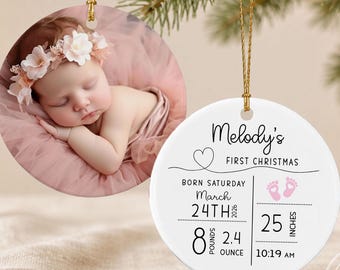 Könnte beinhalten: Zwei runde Keramikornamente mit goldenen Aufhängeschnüren. Eines zeigt ein Foto eines schlafenden Säuglings mit Blumenstirnband. Das andere ist weiß mit schwarzem Text Melody's First Christmas, Geburtsdaten und rosa Fußabdrücken.