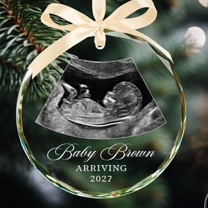 May include: A circular clear glass ornament featuring a black and white ultrasound image of a fetus. The text reads Baby Brown, ARRIVING, and 2027. The ornament is suspended by a light beige ribbon tied in a bow at the top.
