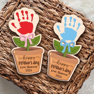 Puede incluir: Dos regalos de madera con forma de flor para el Día de la Madre. Uno tiene una huella de mano roja, el otro una huella de mano azul. Cada uno tiene una maceta con el texto "Happy Mother's Day, Love Shianne 2026". Lazos rosas y azules adornan los tallos.