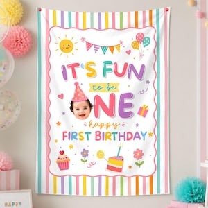 May include: A rectangular birthday backdrop featuring a white center with colorful illustrations of a sun, balloons, a cupcake, a cake, and flowers. The text reads IT'S FUN to be ONE happy FIRST BIRTHDAY with a photo of a baby wearing a party hat.