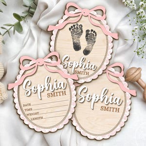May include: Wooden baby announcement plaques with pink bows and scalloped edges. The plaques feature the name "Sophia Smith" in script, with one showing footprints and others with space for birth details. A crocheted bunny and wooden rattle are also present.