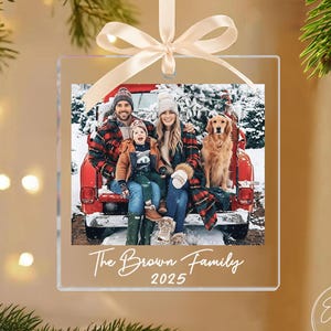 May include: Clear square Christmas ornament with a family photo inside, featuring a family and a dog in a red truck bed. The ornament has a cream-colored ribbon for hanging and the text "The Brown Family 2025".