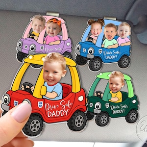 May include: A collection of colorful car-shaped visor clips, each featuring a child's face. The cars are in red, green, blue, and purple, with the text "Drive Safe DADDY!" on each. The clips are designed to attach to a car visor.