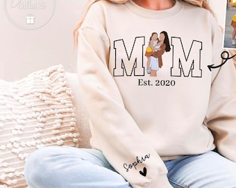 Op de afbeelding: Een beige sweatshirt met de tekst "MOM" in zwarte letters en de tekst "Est. 2020" eronder. Het sweatshirt heeft een klein hartje met de naam "Sophia" op de mouw. Het sweatshirt wordt gedragen door een persoon. De afbeelding bevat ook een kleinere afbeelding van twee meisjes.