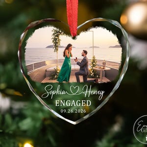 Puede incluir: Un adorno de cristal en forma de coraz&oacute;n con una fotograf&iacute;a impresa de una propuesta de matrimonio en la cubierta de un barco. El texto dice Sophia y Henry, ENGAGED, y 09.26.2026. Una cinta roja est&aacute; sujeta arriba para colgarlo del &aacute;rbol.