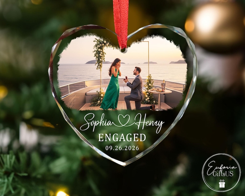 Puede incluir: Un adorno de cristal en forma de coraz&oacute;n con una fotograf&iacute;a impresa de una propuesta de matrimonio en la cubierta de un barco. El texto dice Sophia y Henry, ENGAGED, y 09.26.2026. Una cinta roja est&aacute; sujeta arriba para colgarlo del &aacute;rbol.