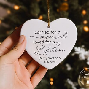 May include: White heart-shaped ornament with the text "carried for a moment loved for a lifetime" and the name "Baby Watson" and the date "5.15.2024".