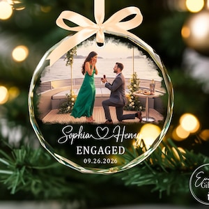 Puede incluir: Un adorno navide&ntilde;o circular de vidrio con una fotograf&iacute;a impresa de una propuesta de matrimonio en un barco. El texto dice Sophia y Henry, ENGAGED, y 09.26.2026. Una cinta de sat&eacute;n color crema est&aacute; atada en la parte superior para colgarlo.