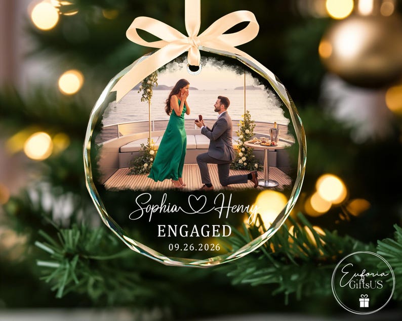 Puede incluir: Un adorno navide&ntilde;o circular de vidrio con una fotograf&iacute;a impresa de una propuesta de matrimonio en un barco. El texto dice Sophia y Henry, ENGAGED, y 09.26.2026. Una cinta de sat&eacute;n color crema est&aacute; atada en la parte superior para colgarlo.