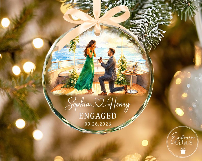 Puede incluir: Un adorno navide&ntilde;o de cristal redondo y facetado con una ilustraci&oacute;n impresa de una propuesta de matrimonio en un barco. El texto dice Sophia, un icono de coraz&oacute;n, Henry, ENGAGED y 09.26.2026. Una cinta beige claro est&aacute; atada en la parte superior.