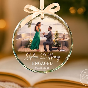 Puede incluir: Un adorno de cristal circular con una fotograf&iacute;a impresa de una propuesta de matrimonio en un barco. El texto dice Sophia Henry, ENGAGED y 09.26.2026. El adorno est&aacute; rematado con un lazo de sat&eacute;n de color crema para colgarlo.