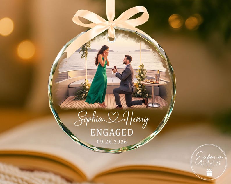 Puede incluir: Un adorno de cristal circular con una fotograf&iacute;a impresa de una propuesta de matrimonio en un barco. El texto dice Sophia Henry, ENGAGED y 09.26.2026. El adorno est&aacute; rematado con un lazo de sat&eacute;n de color crema para colgarlo.