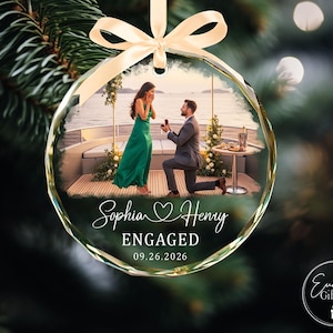 Puede incluir: Un adorno navide&ntilde;o redondo de cristal transparente con una fotograf&iacute;a impresa de una propuesta de matrimonio en un barco. El texto dice Sophia y Henry, ENGAGED, y 09.26.2026. Una cinta de sat&eacute;n color crema est&aacute; atada arriba para colgarlo.