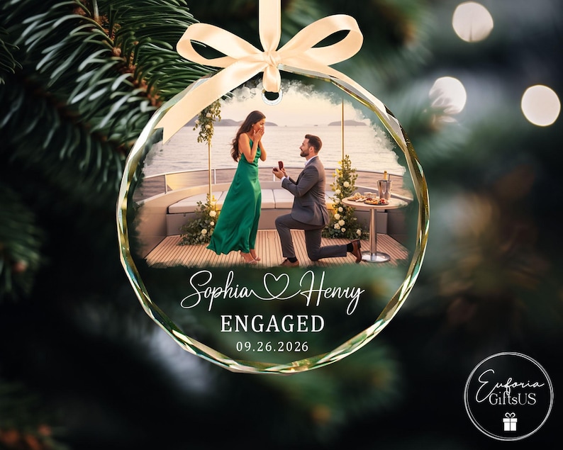 Puede incluir: Un adorno navide&ntilde;o redondo de cristal transparente con una fotograf&iacute;a impresa de una propuesta de matrimonio en un barco. El texto dice Sophia y Henry, ENGAGED, y 09.26.2026. Una cinta de sat&eacute;n color crema est&aacute; atada arriba para colgarlo.