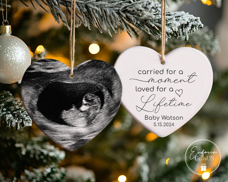 May include: Two white heart-shaped Christmas ornaments. One ornament features a black and white ultrasound image of a baby. The other ornament has the text "carried for a moment loved for a lifetime" with the name "Baby Watson" and the date "5.15.2024".