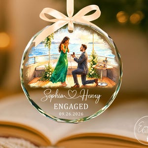Puede incluir: Un adorno de cristal redondo y facetado con una ilustraci&oacute;n impresa de una propuesta de matrimonio en un barco. El texto dice Sophia, un s&iacute;mbolo de coraz&oacute;n, Henry, ENGAGED y 09.26.2026. Una cinta de sat&eacute;n color crema est&aacute; atada en la parte superior.