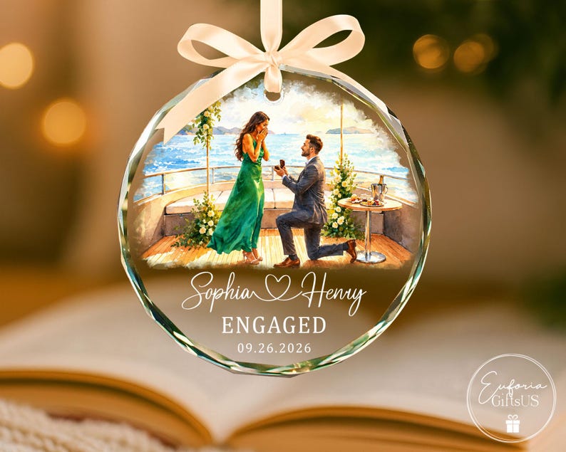 Puede incluir: Un adorno de cristal redondo y facetado con una ilustraci&oacute;n impresa de una propuesta de matrimonio en un barco. El texto dice Sophia, un s&iacute;mbolo de coraz&oacute;n, Henry, ENGAGED y 09.26.2026. Una cinta de sat&eacute;n color crema est&aacute; atada en la parte superior.
