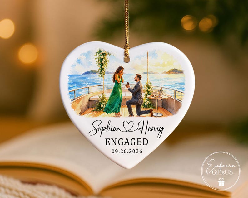 Puede incluir: Un adorno de cer&aacute;mica blanca en forma de coraz&oacute;n con una ilustraci&oacute;n en acuarela de una propuesta de matrimonio en la cubierta de un barco. El texto dice Sophia, un icono de coraz&oacute;n, Henry, ENGAGED y 09.26.2026. Cuelga de un cord&oacute;n dorado.