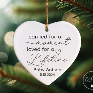 May include: White heart-shaped ornament with the text "carried for a moment loved for a lifetime" and the name "Baby Watson" and the date "5.15.2024".