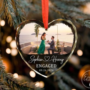 Puede incluir: Un adorno de cristal en forma de coraz&oacute;n con una fotograf&iacute;a impresa de una propuesta de matrimonio en la cubierta de un barco. El texto dice Sophia y Henry, ENGAGED, y 09.26.2026. El adorno cuelga de una cinta roja sobre un fondo desenfocado.
