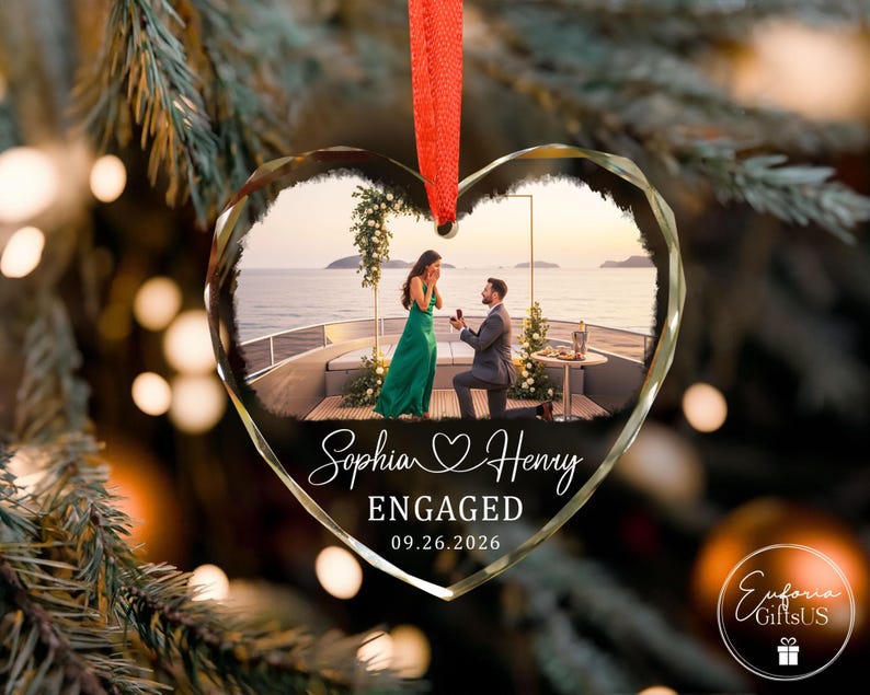 Puede incluir: Un adorno de cristal en forma de coraz&oacute;n con una fotograf&iacute;a impresa de una propuesta de matrimonio en la cubierta de un barco. El texto dice Sophia y Henry, ENGAGED, y 09.26.2026. El adorno cuelga de una cinta roja sobre un fondo desenfocado.