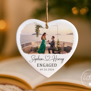 Puede incluir: Un adorno de cer&aacute;mica blanco en forma de coraz&oacute;n que cuelga de un cord&oacute;n dorado. El centro presenta una fotograf&iacute;a impresa de una propuesta de matrimonio en un barco. Debajo de la foto, el texto dice Sophia and Henry, ENGAGED, y la fecha 09.26.2026.